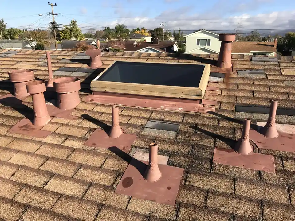 The same roof before: painted galvanized pipes and vents in rust brown on tan shingles around a skylight