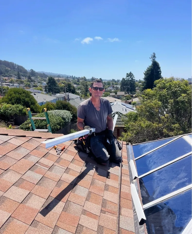 Adam, lead roofer at Sonrise Roofing, kneeling beside a skylight on a composition shingle roof with the East Bay hills behind him