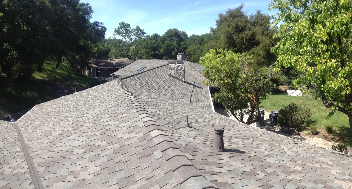 Long view down a new gray-brown shingle ridge with a stone-coated vent and pipe, oaks and a lawn below