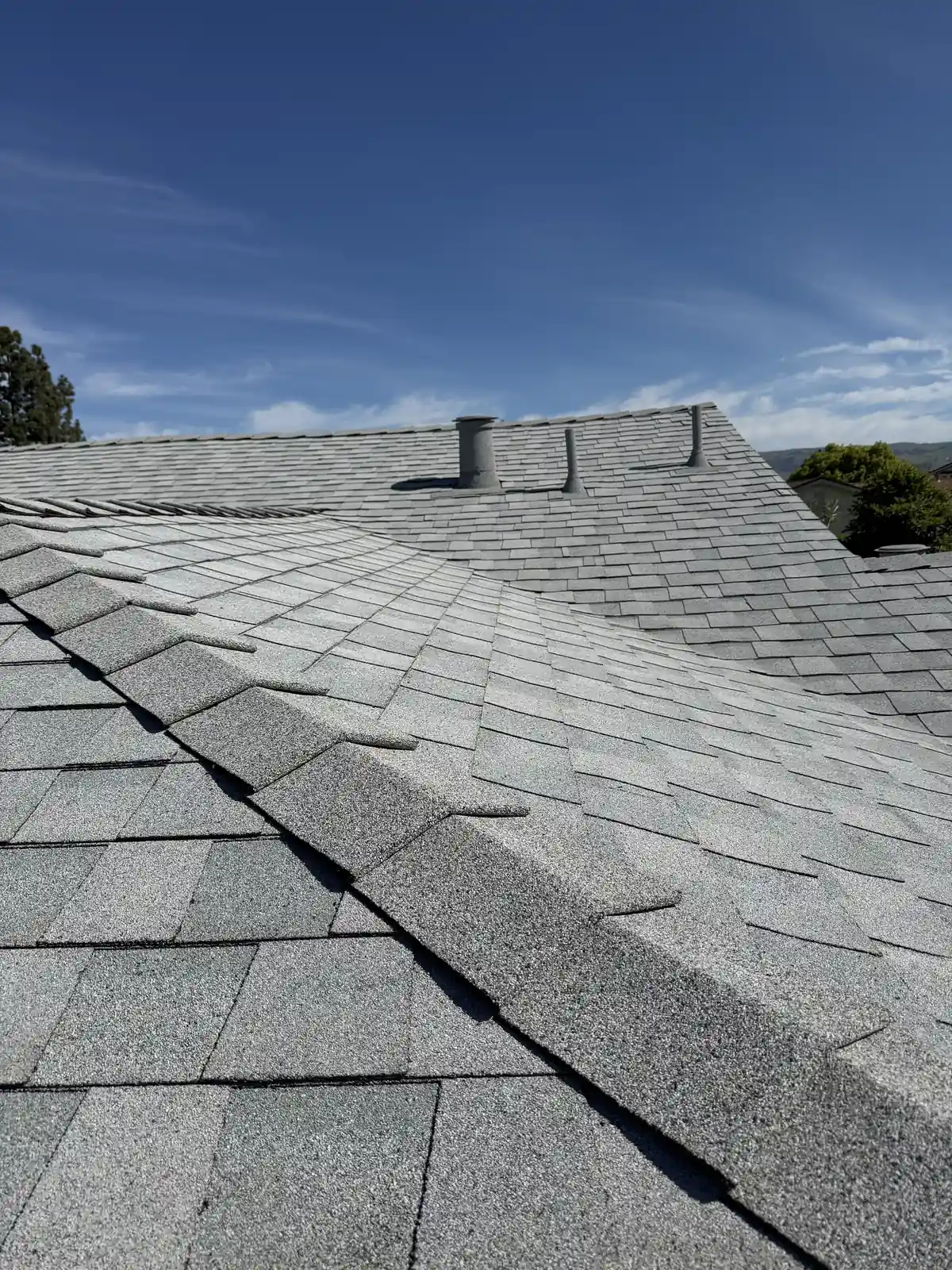 Light gray shingle roof with a stone-coated ridge vent line and a stone-coated pipe near the ridge under a wide sky
