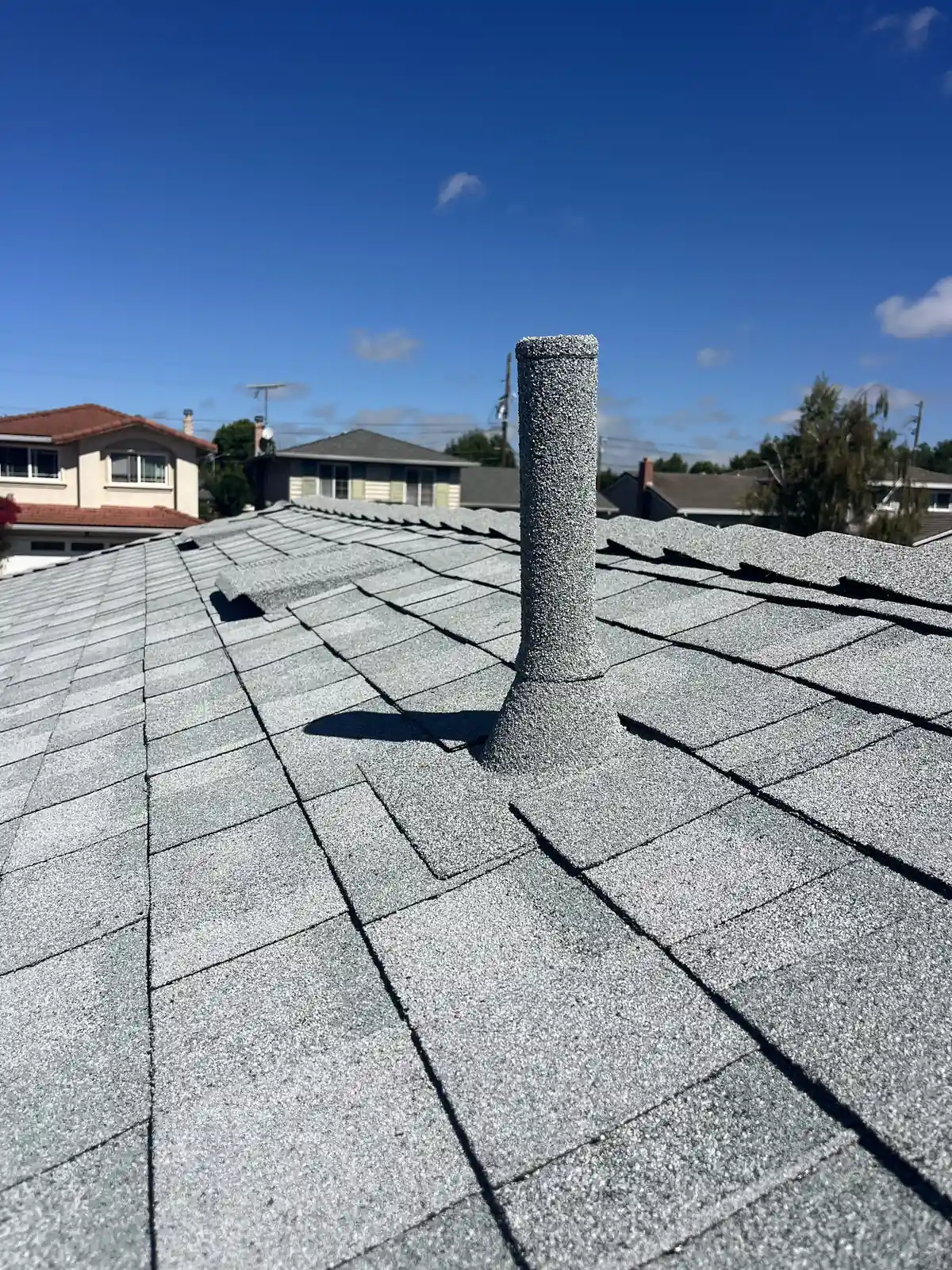Tall stone-coated pipe sleeve on a new gray shingle roof under a blue sky, neighboring rooftops behind